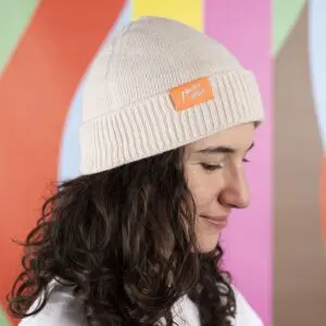 Une personne aux longs cheveux bruns bouclés porte un bonnet Bonnet beige, Montreux Jazz Festival, de couleur crème avec une étiquette orange. L'arrière-plan présente des rayures diagonales colorées en vert, orange, rouge, rose et marron.