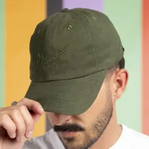 Un homme en chemise blanche et moustache abaisse le bord d'une casquette Casquette Montreux Jazz, Forest, couvrant partiellement son visage. La casquette vert olive comporte un texte brodé. Le fond présente des rayures verticales de couleur pastel.