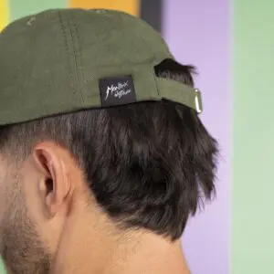 A close-up shows the back of a person's head wearing the Cap Montreux Jazz, Forest. The olive green cap features an adjustable strap with a black label and white handwriting-style text. The colorful background is blurred.