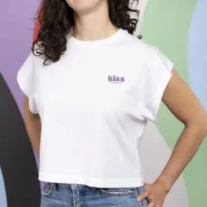 A woman with wavy brown hair wears the "T-shirt Crop, Kiss"—a white crop top with "kiss" in purple letters—posing with her hand on her hip before a colorful abstract background in Montreux.