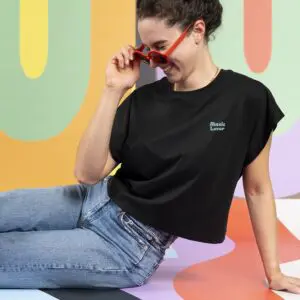 A woman with dark hair in a bun, wearing red sunglasses and blue jeans, smiles on a colorful abstract background. She is dressed casually in the "T-shirt Crop, Music Lover.