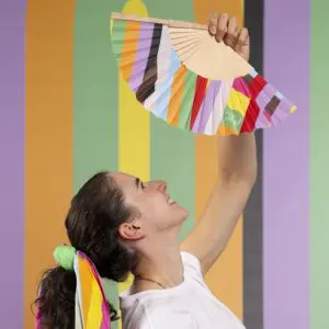 Une personne aux cheveux longs attachés avec un foulard rayé arc-en-ciel brandit un éventail à main Éventail Lakwena sur un fond rayé multicolore et vibrant au festival de Montreux.