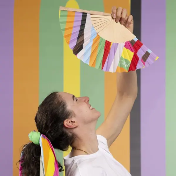 Une personne aux cheveux longs attachés avec un foulard rayé arc-en-ciel brandit un éventail à main Éventail Lakwena sur un fond rayé multicolore et vibrant au festival de Montreux.