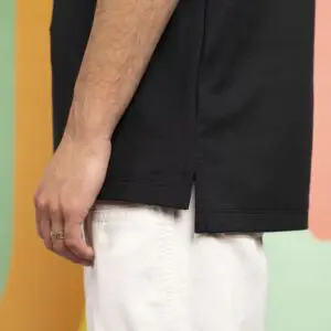A person wearing a black short-sleeve shirt and white pants stands against a colorful background. Only the side of their torso and arm are visible. The person has a gold ring on their finger.