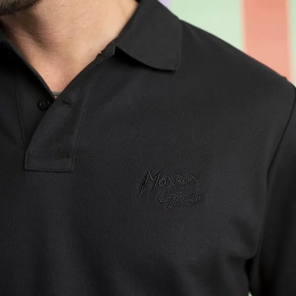 Close-up of a person wearing a black polo shirt with Montreux Edition embroidered in black on the chest, standing in front of a pastel-colored background. The persons head is partially out of the frame.
