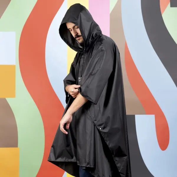 Wearing the Rain Poncho Durable with a hood, a person stands before a vivid, abstract backdrop of bold letters inspired by the Montreux Festival. They adjust their sleeve and glance down.