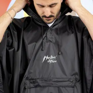 A person adjusts the hood on a black Rain Poncho Durable, featuring a white iconic logo on the front pocket. Their face is partly visible against a backdrop of blurred, colorful shapes.