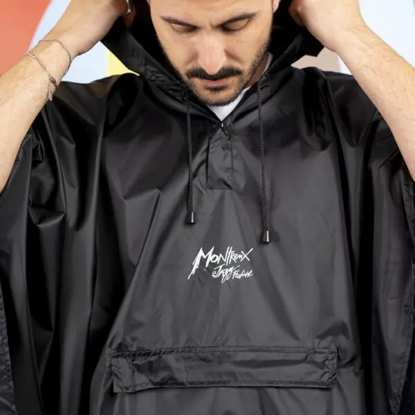 A person adjusts the hood on a black Rain Poncho Durable, featuring a white iconic logo on the front pocket. Their face is partly visible against a backdrop of blurred, colorful shapes.