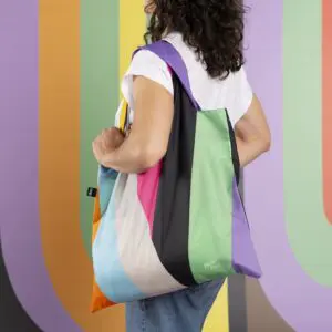 A person with wavy dark hair, dressed in a white shirt and jeans, holds the Bag LOQI Lakwena tote. The backdrop’s bold vertical stripes in pastel and bright hues echo the bag’s Montreux Jazz-inspired design.