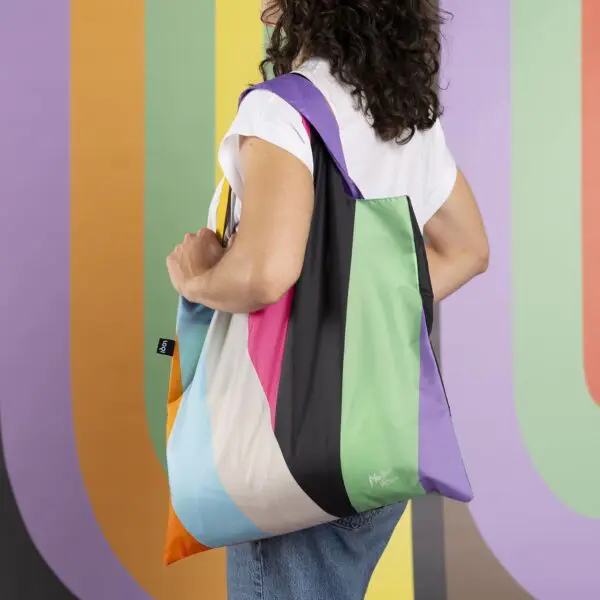 A person with wavy dark hair, dressed in a white shirt and jeans, holds the Bag LOQI Lakwena tote. The backdrop’s bold vertical stripes in pastel and bright hues echo the bag’s Montreux Jazz-inspired design.