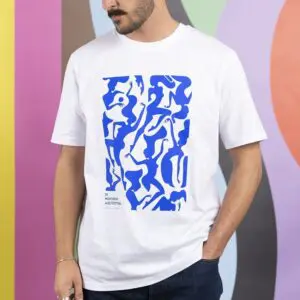A man wears the 2017 T-Shirt Malika Favre in blue, featuring an abstract design and small “57e Montreux Jazz Festival” text, as he stands against a colorful wavy-patterned wall.