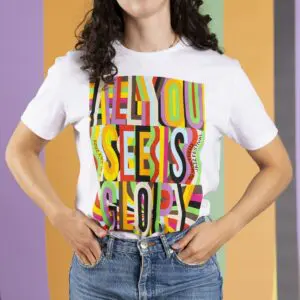 Une personne aux cheveux bruns bouclés portant un T-shirt blanc avec un texte gras et coloré indiquant ALL YOU SEE IS GLORY, debout devant un fond rayé pastel.