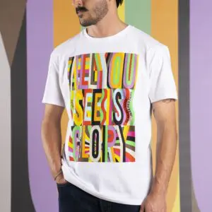 Un homme vêtu d'un T-shirt blanc avec des lettres colorées et audacieuses indiquant ALL YOU SEE IS GLORY, debout devant un fond rayé et multicolore.