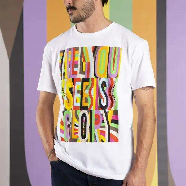 Un homme vêtu d'un T-shirt blanc avec des lettres colorées et audacieuses indiquant ALL YOU SEE IS GLORY, debout devant un fond rayé et multicolore.