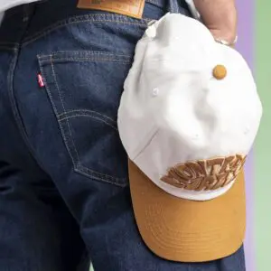 A person in dark blue jeans has a Vintage Cap Montreux Jazz, Camel, hooked to their back pocket, set against a softly blended green and purple background.