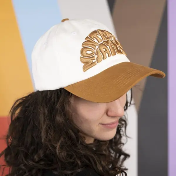 Person with long curly hair wearing a white and tan cap that says Montara Falls, standing in front of a colorful geometric background.