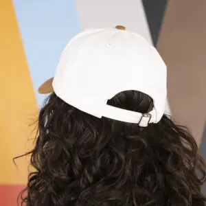 A person with long, curly brown hair is wearing a Vintage Cap Montreux Jazz, Camel, backwards. The background features vertical panels in orange, blue, gray, black, and brown.