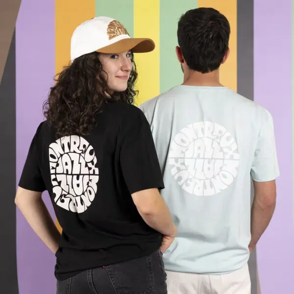 Two people pose before a vibrant striped backdrop, modeling Vintage Montreux Jazz Festival T-Shirts. One wears the black version and a white cap, while the other sports a light blue tee. Both shirts feature bold circular text designs on the back.