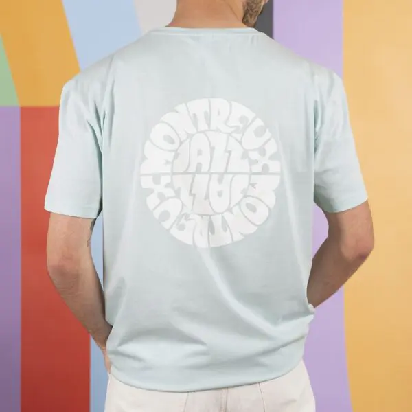 Someone stands with their back to the camera, wearing the T-Shirt Vintage Montreux Jazz Festival, Bleu ciel, featuring a circular white "Montreux Jazz" logo, against a vibrant, geometric background.