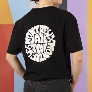 Someone stands against a colorful geometric background wearing the T-Shirt Vintage Montreux Jazz Festival, Black, which features a bold white circular graphic of Montreux Jazz on the back.