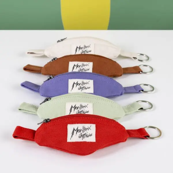 Five Mini Banana Bag Keyrings in cream, brown, purple, green, and red—with zippers, keyrings, and a white Monttrex Signature label—are arranged diagonally, reflecting the playful vibe of this unique accessory.