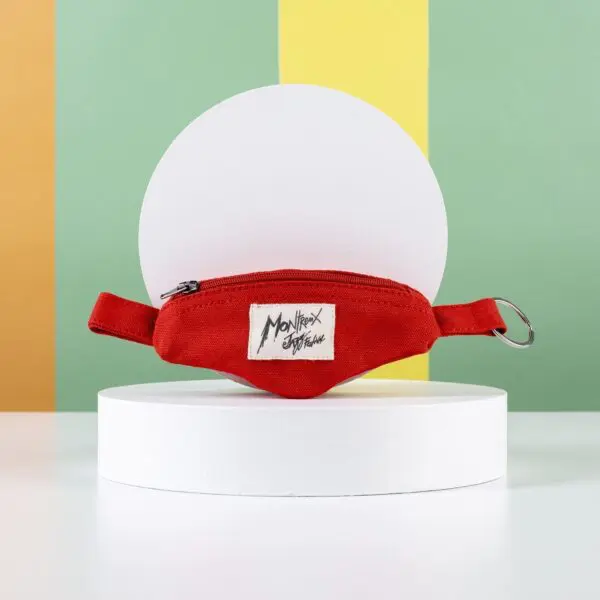 The Mini Banana Bag Keyring, a small red waist bag with a Monthex Stefanie fabric label and Porte-clé Banana Montreux Jazz accent, is displayed on a white round pedestal against vibrant green, yellow, and orange stripes.