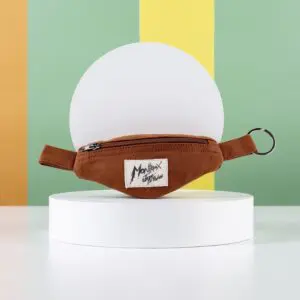 The Mini Banana Bag Keyring, a small brown bag with a zipper and Montreux Jazz label, is displayed on a white circular stand against a vibrant green, yellow, and orange striped background.