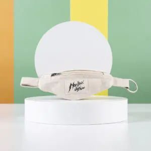 A beige fabric waist bag with a black zipper and a "Monkxfy Artisan" patch is displayed on a round white pedestal, accented by the playful Mini Banana Bag Keyring, set against orange, yellow, and green vertical stripes.