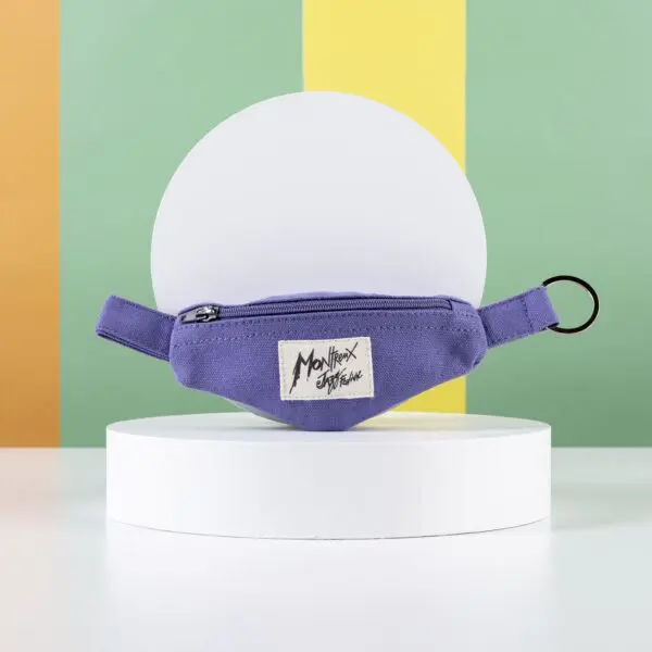 A small purple Mini Banana Bag Keyring with a Monhexx Stylehouse fabric label rests on a white pedestal, set against vibrant orange, yellow, and green stripes—a playful accent for your Banana Montreux Jazz accessories.
