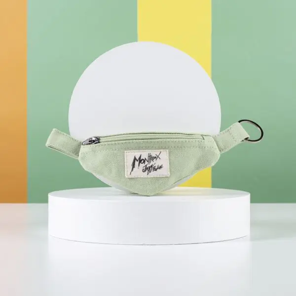 A Mini Banana Bag Keyring—a small light green zippered fabric pouch with Monhex Studio label—styled like a porte-clé Banana Montreux Jazz, shown on a white circular stand with a green, yellow, and orange striped background.