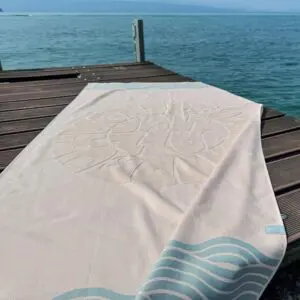 The Beach Towel Coral, Vintage, with its light color and decorative pattern, is spread out on a wooden dock beside calm blue water, with the dock and a distant horizon in the background.