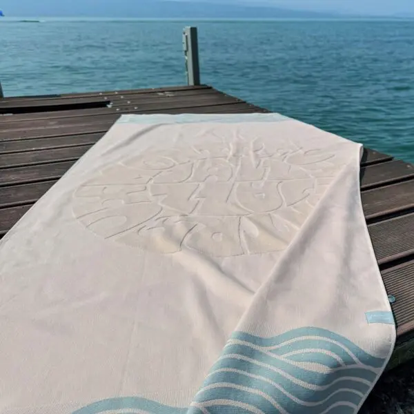 The Beach Towel Coral, Vintage, with its light color and decorative pattern, is spread out on a wooden dock beside calm blue water, with the dock and a distant horizon in the background.
