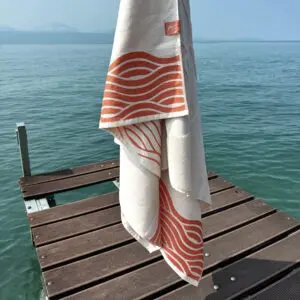 The Beach Towel Coral, Vintage, in cream with orange coral wave patterns, hangs on a wooden dock above clear, blue-green water, with a calm lake and Montreux’s hazy mountains in the background.
