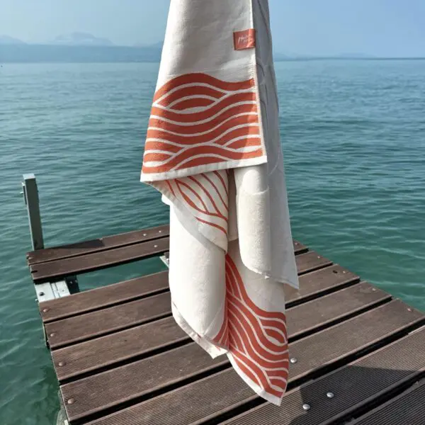 The Beach Towel Coral, Vintage, in cream with orange coral wave patterns, hangs on a wooden dock above clear, blue-green water, with a calm lake and Montreux’s hazy mountains in the background.