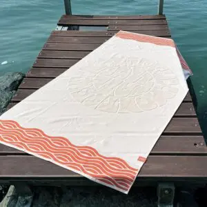 The Beach Towel Coral, Vintage features cream fabric with orange wavy stripes on the edges and a subtle circular center design, perfect for relaxing on a wooden dock above turquoise water.