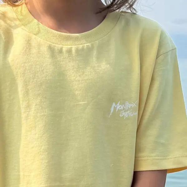 A child wearing a pale yellow T-Shirt Fun, Kids featuring the Montreux festival’s white logo on the chest; image shows neck to mid-torso with long hair partially visible.