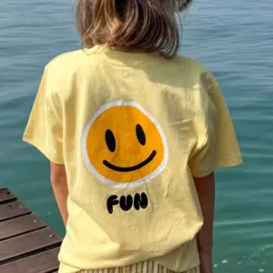 Une personne aux cheveux mi-longs se tient au bord de l'eau à Montreux, vêtue d'un T-Shirt jaune Fun, Enfant avec un smiley et "FUN" au dos. Un quai en bois est visible sur la gauche.