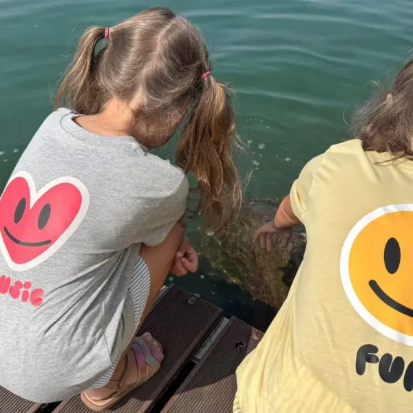 Two young girls in pigtails wear "T-Shirt Fun, Kids" shirts with big smiley faces as they lean over a wooden dock by green water in Montreux, curiously watching something beneath the surface.