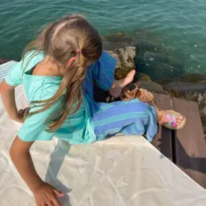 A young girl with light brown hair in a ponytail sits by the water on a mat, wearing a turquoise shirt, striped blue pants, colorful sandals, and glancing down near rocks. The Beach Towel Coral, Vintage is placed next to her.
