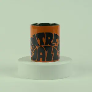 The Mug Vintage Orange, featuring bold black Montreux Jazz-inspired lettering, sits on a round white pedestal against a pale green background.