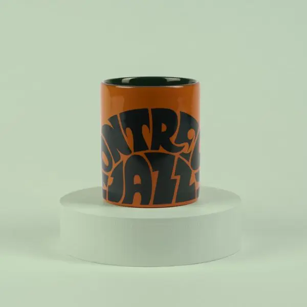 The Mug Vintage Orange, featuring bold black Montreux Jazz-inspired lettering, sits on a round white pedestal against a pale green background.