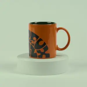 The Mug Vintage Orange features bold black abstract lettering inspired by Montreux Jazz designs. Displayed on a round white pedestal against a pale green background, its interior shows a dark green hue.