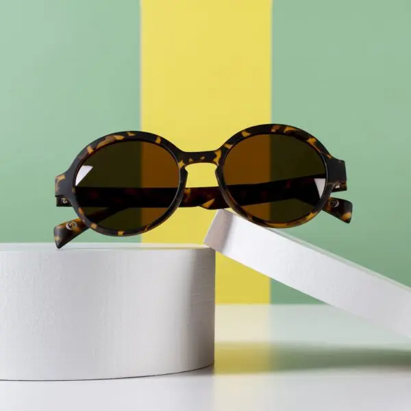 Fly Amsterdam Light Tortoiseshell Glasses with dark lenses, shown on white cylindrical podiums, set against a green and yellow striped background inspired by Montreux Jazz.