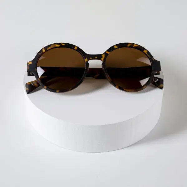 Fly Amsterdam Light Tortoiseshell Glasses with dark lenses are showcased on a white cylindrical platform against a light background.