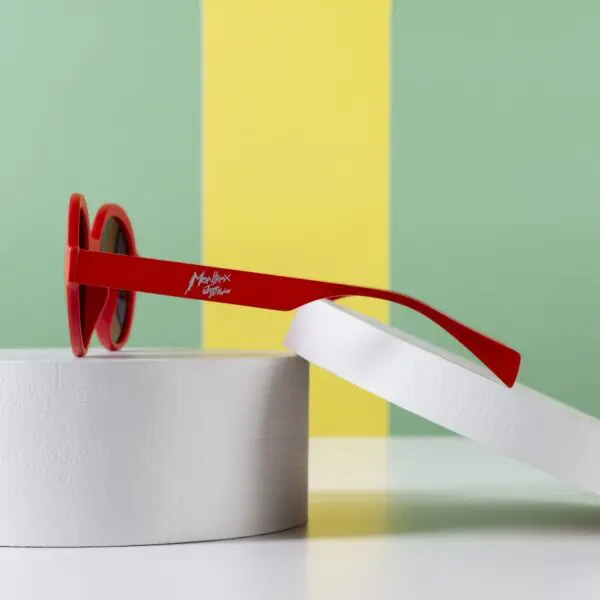 A pair of Fly Amsterdam Peach Glasses with round lenses and a signature on the side rests on two white cylindrical platforms set against a green background with a vertical yellow stripe, evoking the iconic Amsterdam style.
