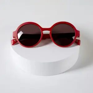 The Fly Amsterdam Peach Glasses, round red sunglasses with dark lenses, are showcased on a white circular platform, reflecting Montreux Jazz vibes and Glasses Amsterdam’s iconic style—all on a plain white background.