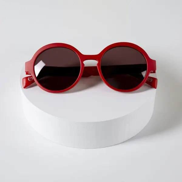 The Fly Amsterdam Peach Glasses, round red sunglasses with dark lenses, are showcased on a white circular platform, reflecting Montreux Jazz vibes and Glasses Amsterdam’s iconic style—all on a plain white background.
