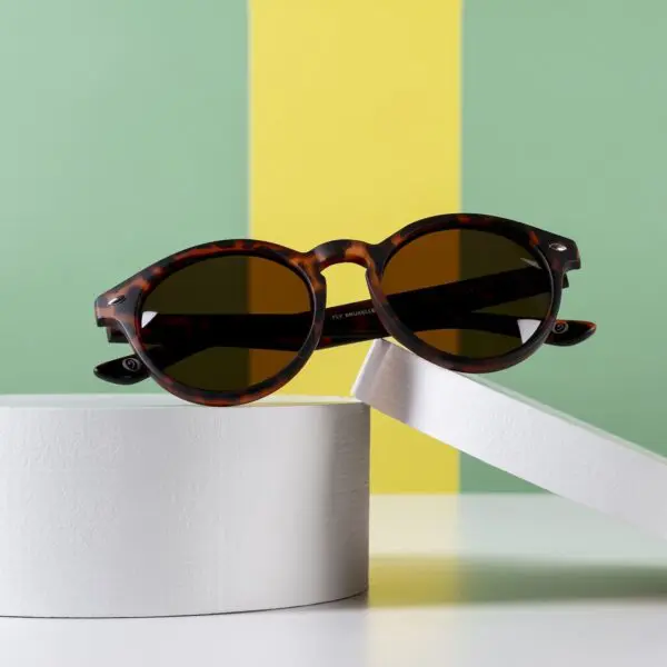 Fly Bruxelles Light Tortoiseshell Glasses with dark lenses, inspired by Montreux Jazz, are showcased on white circular pedestals set against a green and yellow striped background.