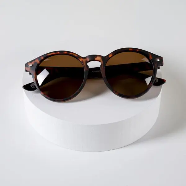 Fly Bruxelles Light Tortoiseshell Glasses with dark lenses rest on a white, circular platform against a plain background—a stylish nod to iconic eyewear and Montreux Jazz festival flair.