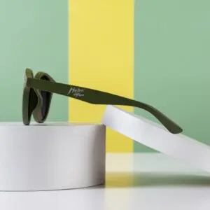 Fly Bruxelles Khaki Glasses displayed on a white circular stand against a green and yellow color-blocked background. The side-on angle highlights the khaki frame and the text on the arm.